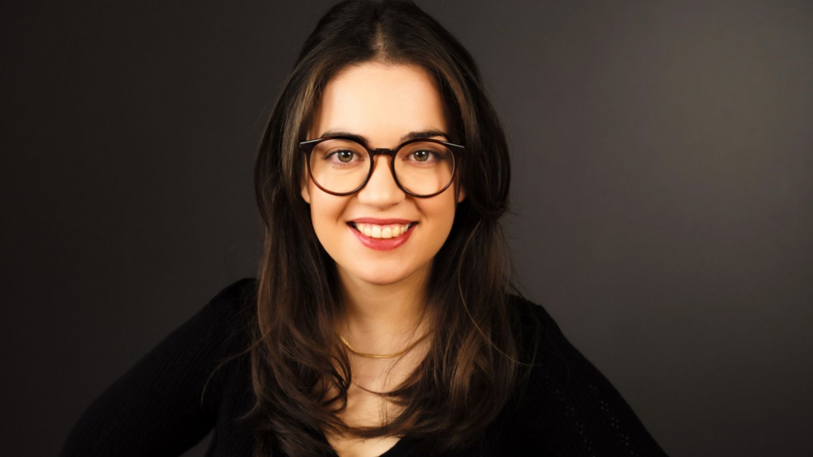 A smiling woman with long dark hair, glasses, and a black top, posed against a dark grey background.